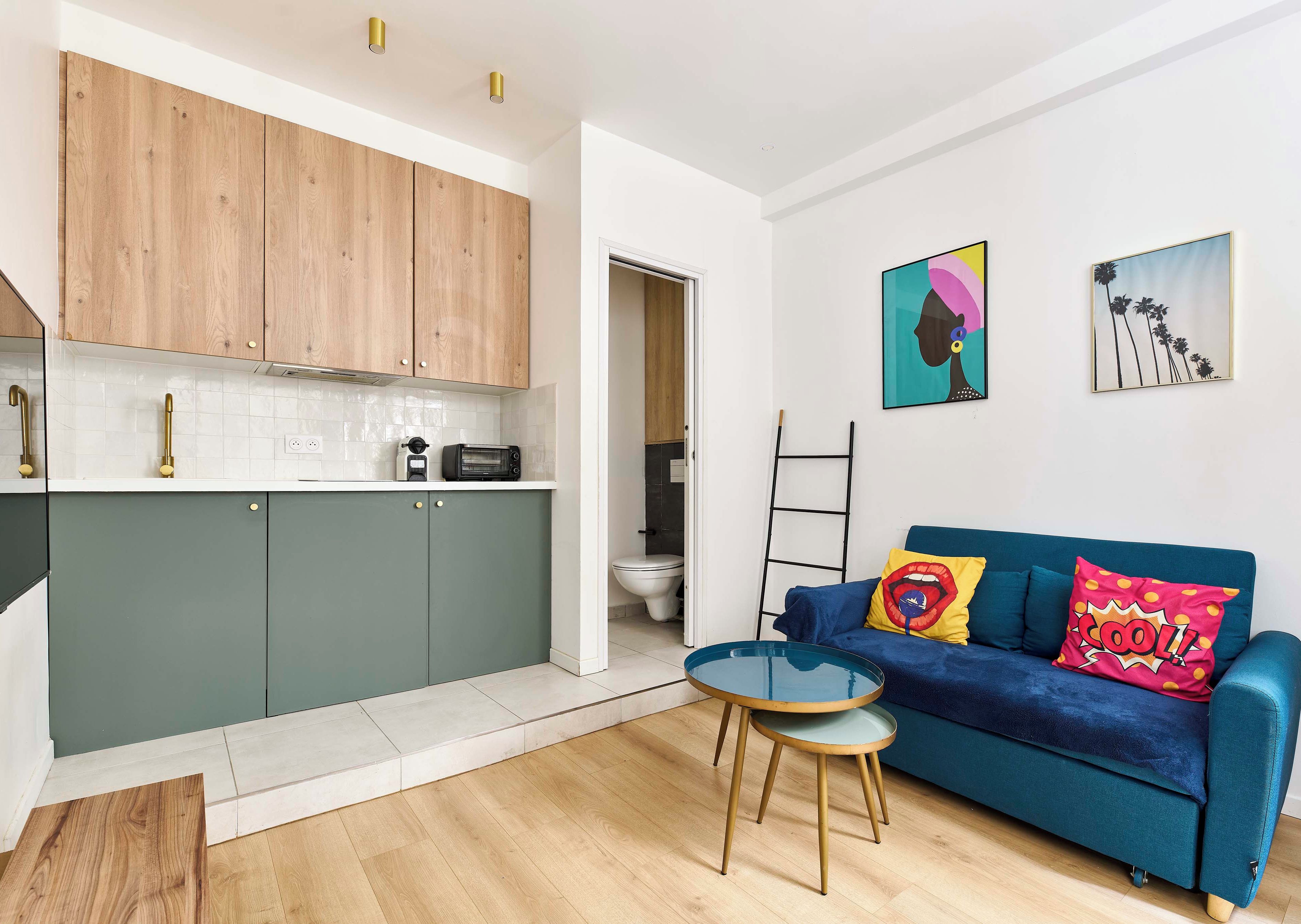 A compact living space with a kitchenette, a blue sofa with colorful cushions, and a small round table.