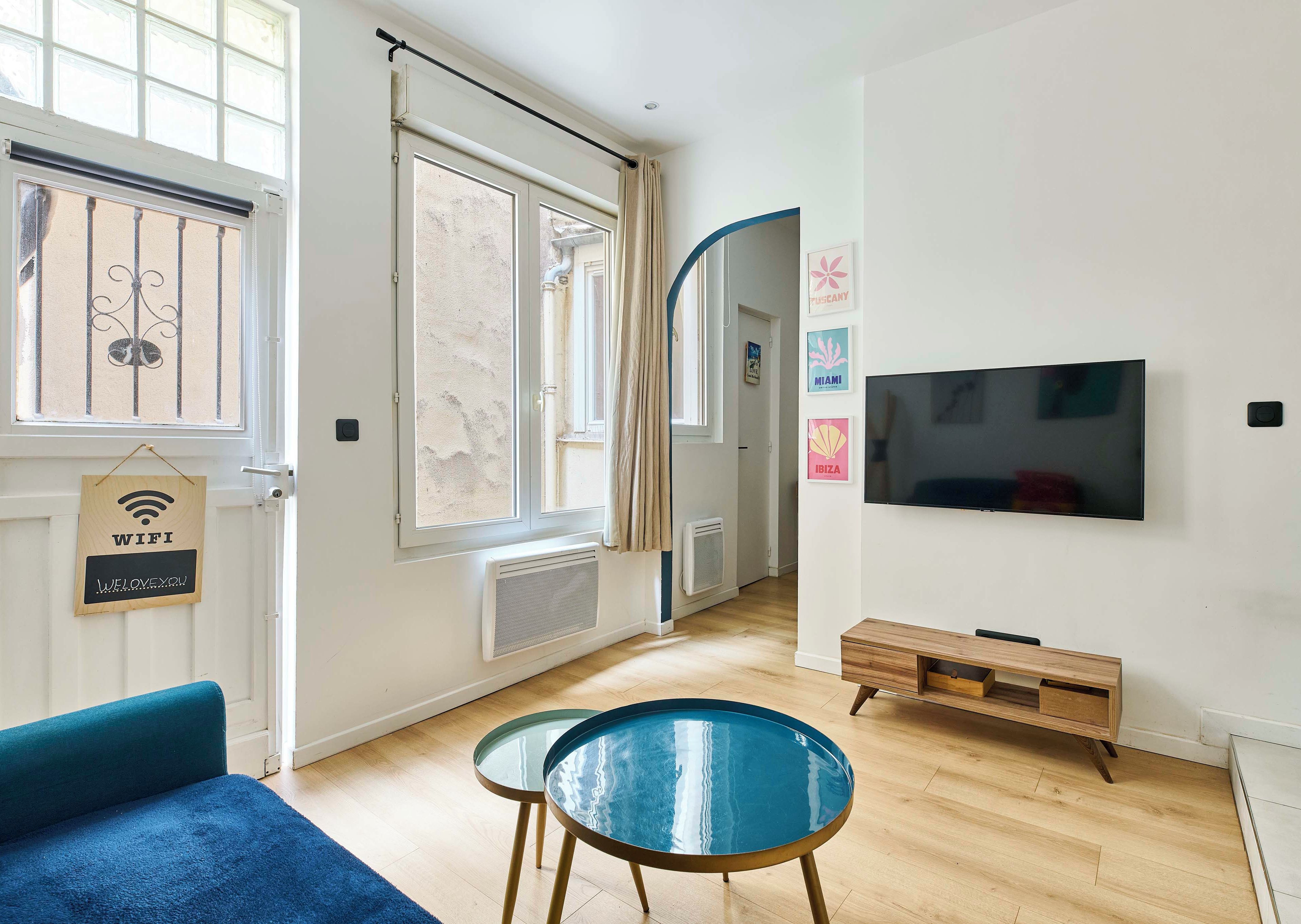 A modern living room with a flat-screen TV, a blue sofa, two glass-top tables, and a sign indicating WIFI availability.