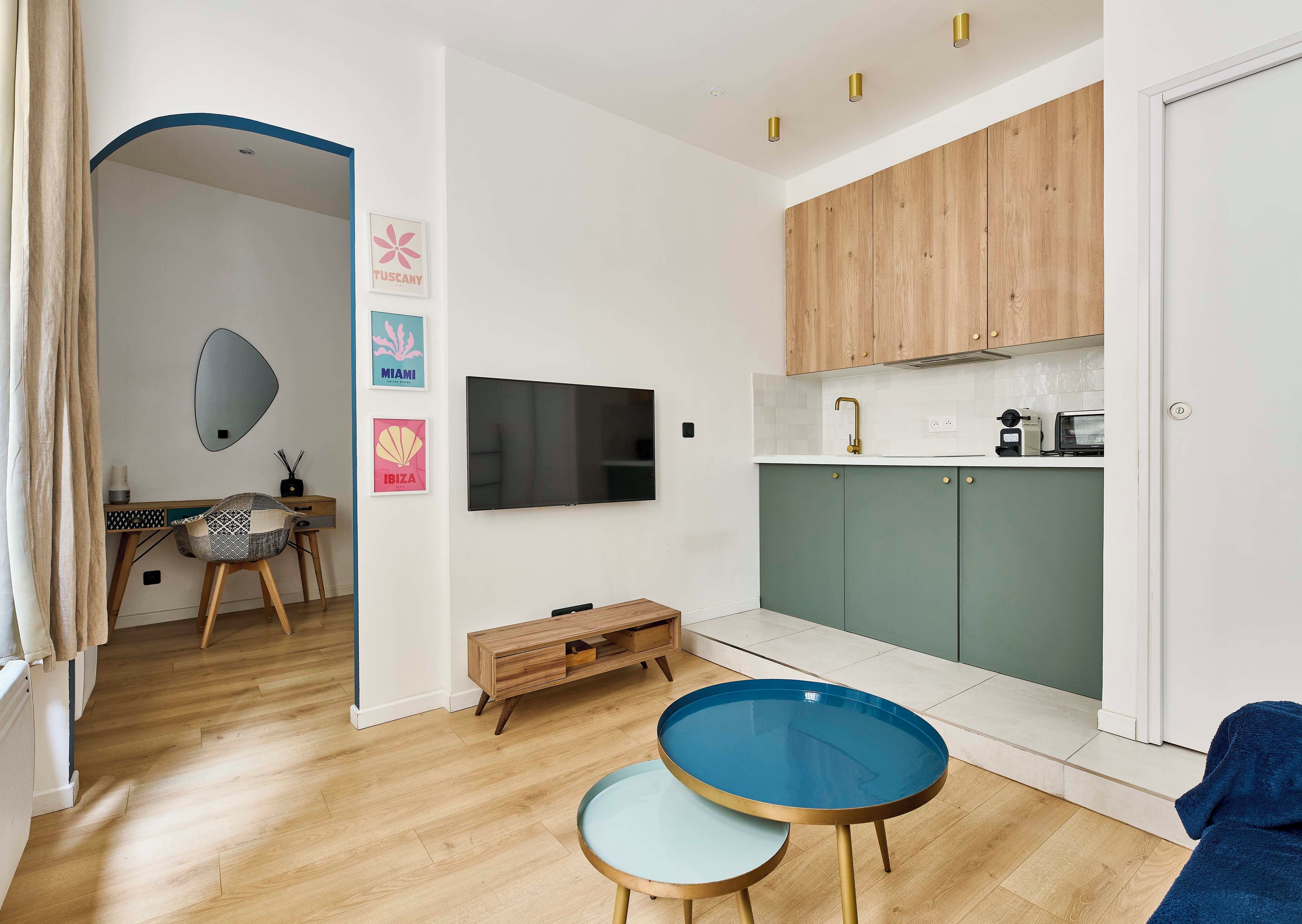 Una sala de estar moderna con piso de madera, un televisor montado en la pared, un comedor con mesa y sillas de madera, y una cocina con gabinetes verdes y encimera blanca.