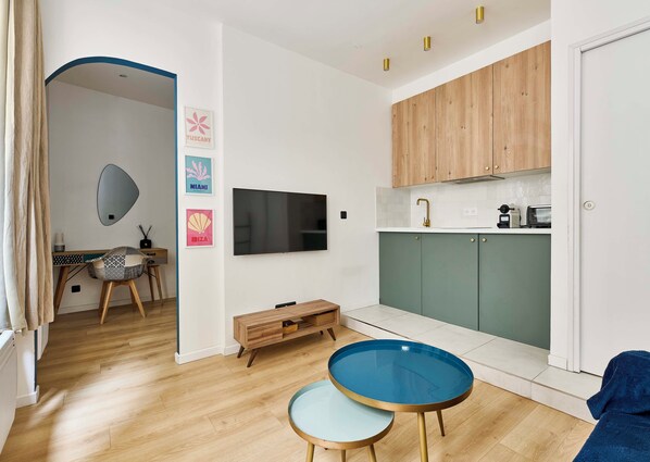 Uma sala de estar moderna com piso de madeira, televisão fixada na parede, área de jantar com mesa e cadeiras de madeira e cozinha com armários verdes e bancada branca.