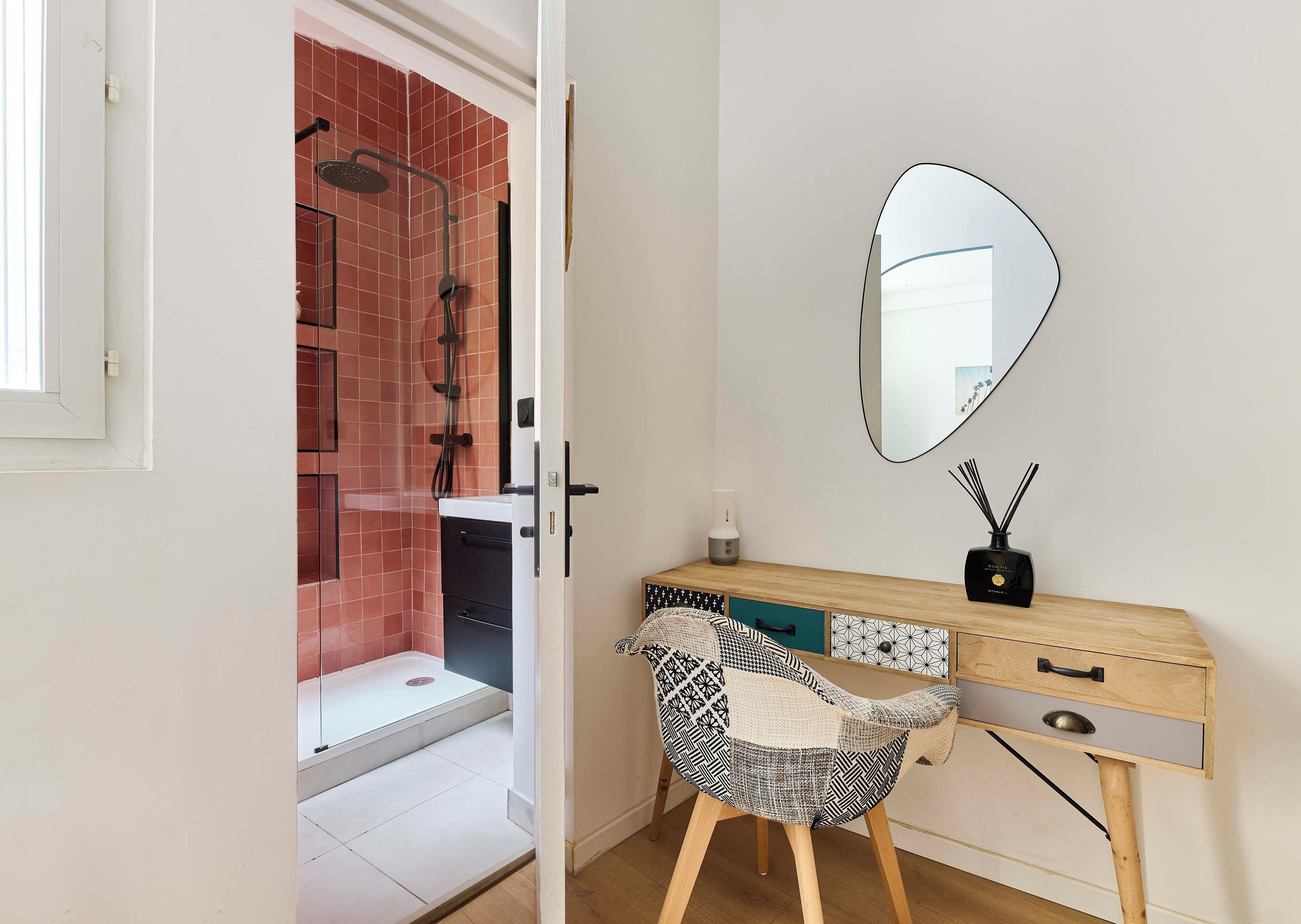 A bathroom with a shower, a mirror, a wooden vanity with a drawer, and a patterned chair.