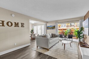 A modern living room with a grey sofa, a coffee table, and a dining area with a small table and chairs. - Beachside Bliss | 1BR | Pool & Patio (Sarasota)