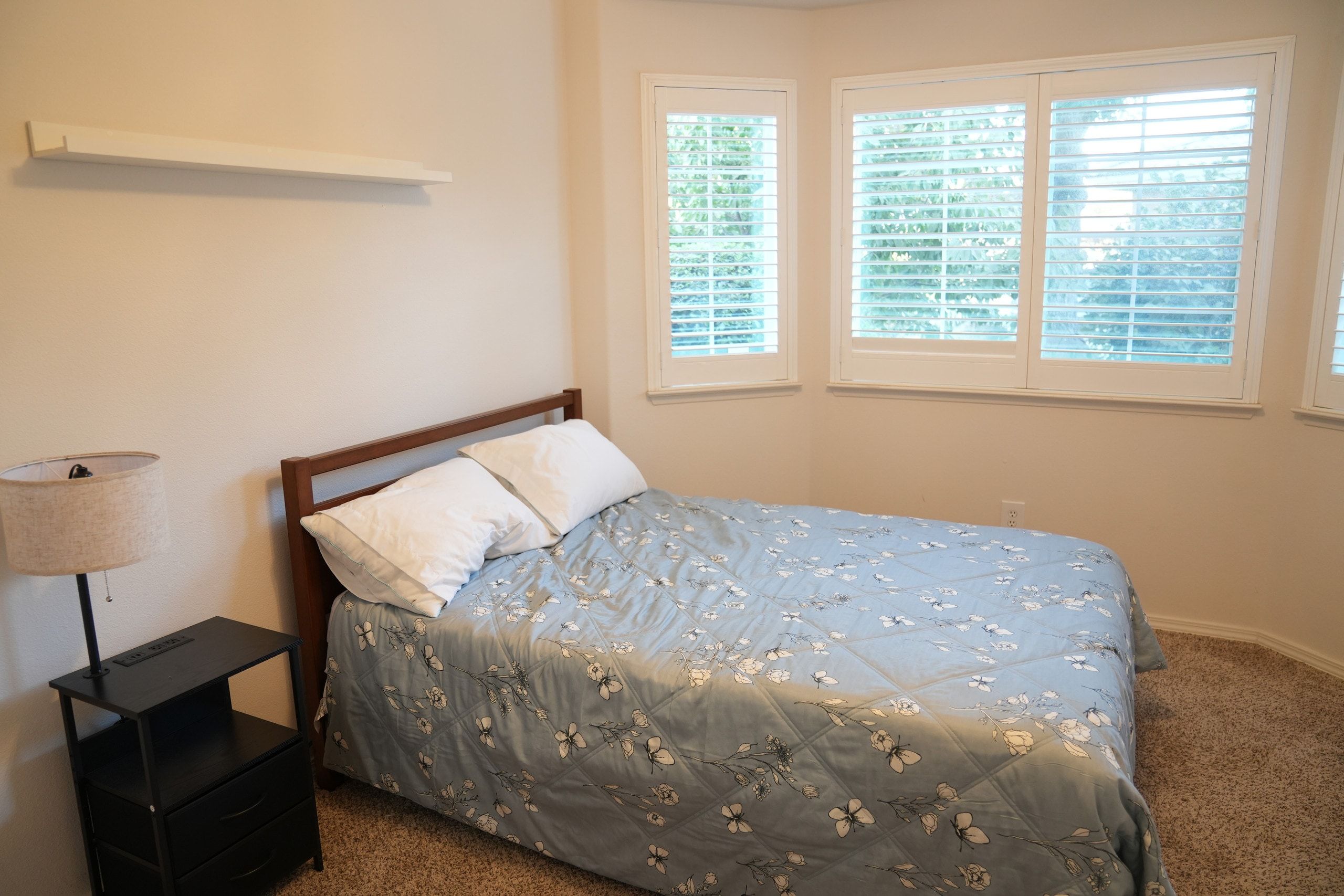 A bedroom with a bed, a nightstand, a lamp, and two windows with blinds.