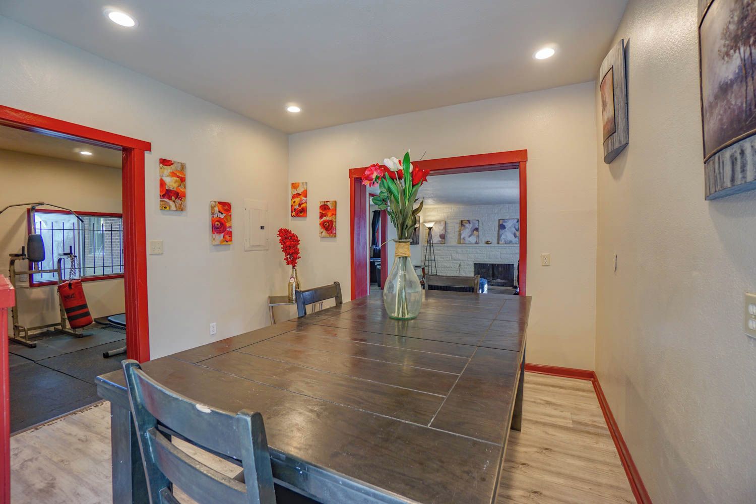 A dining area with a dark wooden table, chairs, a vase with flowers, and a red-framed doorway leading to a kitchen.