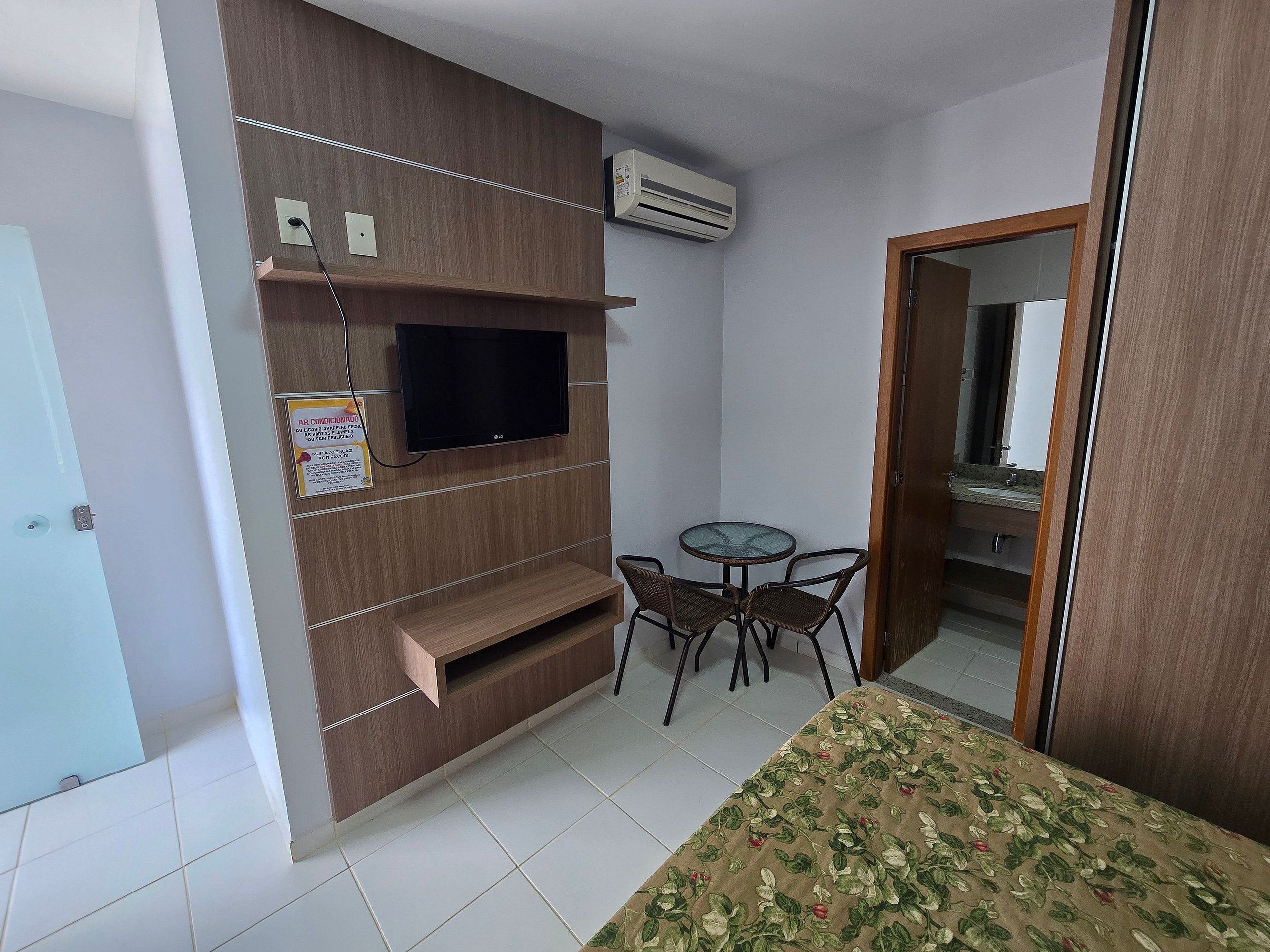 Una habitación pequeña con una televisión instalada en una pared de madera, una mesa con sillas y una cama con colcha estampada.