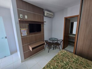 Ein kleines Zimmer mit einem an einer Holzwand befestigten Fernseher, einem Tisch mit Stühlen und einem Bett mit Blumenmusterdecke.