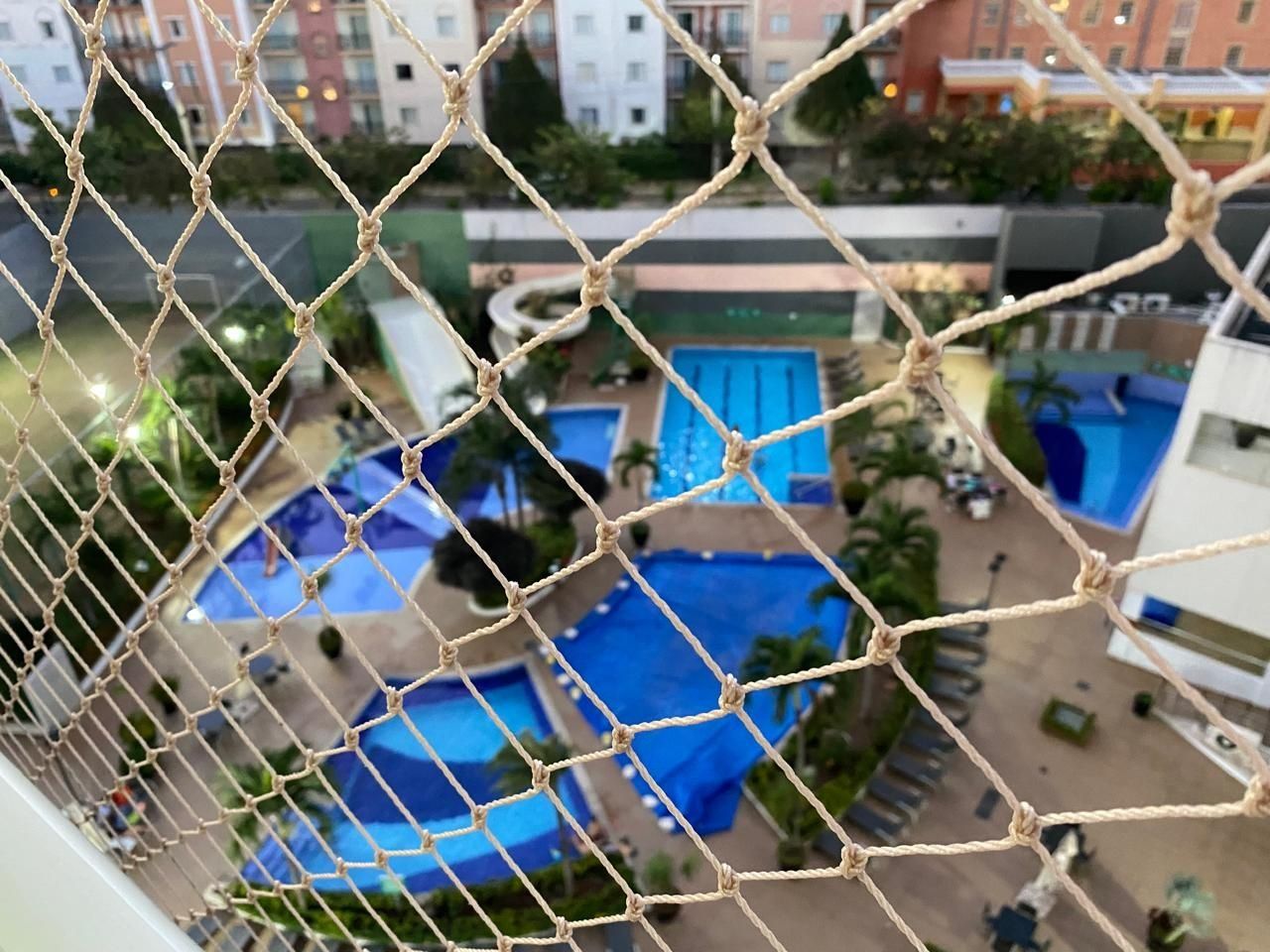 Blick durch ein mit Netz bespanntes Fenster auf ein Hotel mit mehreren Schwimmbecken und einem Parkplatz.