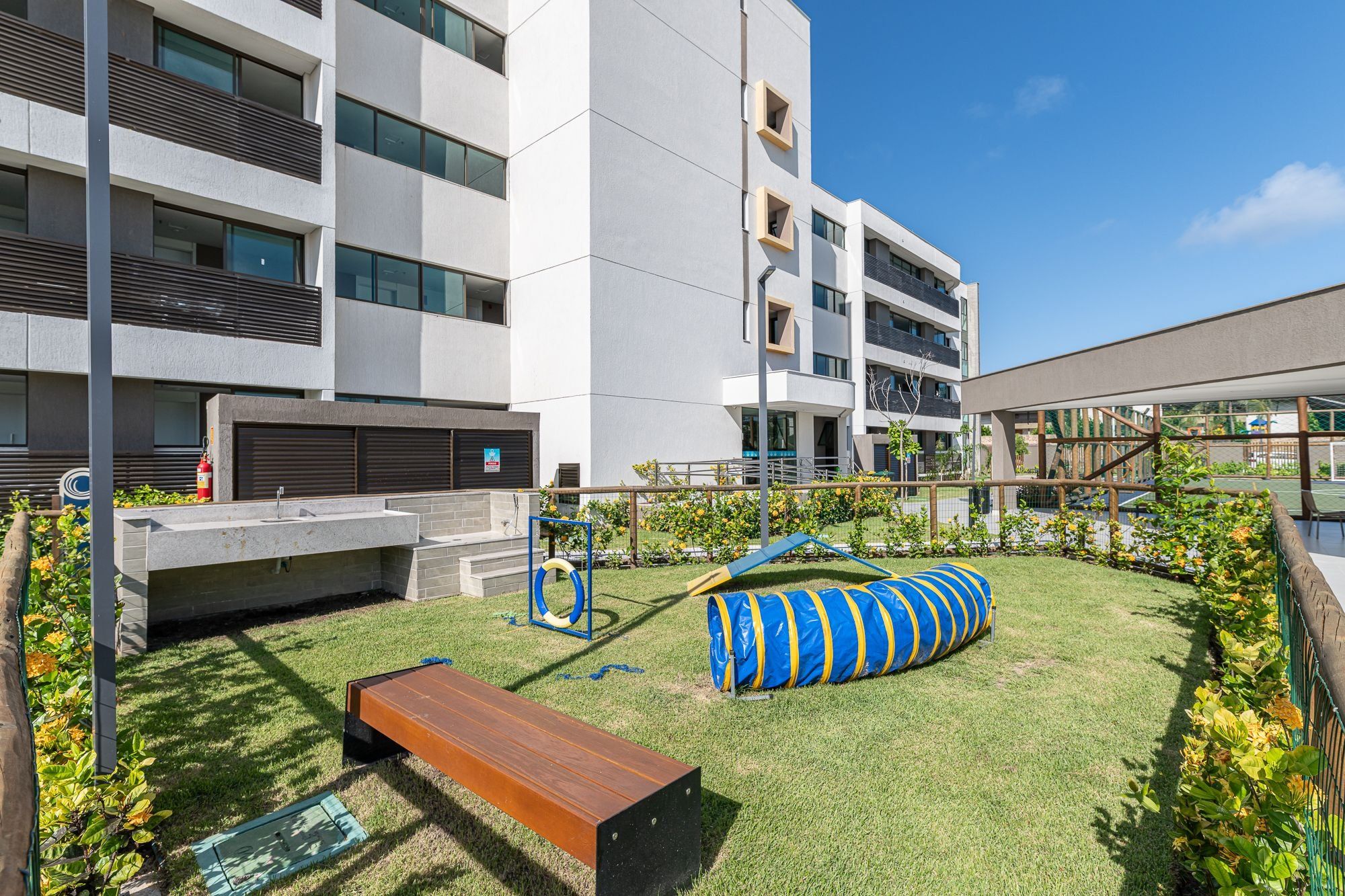 Un edificio moderno con techo plano, un hidrante, un banco y un parque infantil con un tobogán y un túnel azul y amarillo.