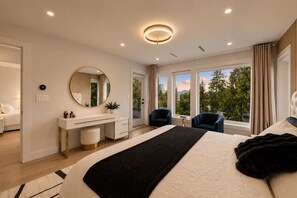 Quarto moderno com uma cama grande, um espelho redondo, uma mesa de cabeceira, uma escrivaninha, duas cadeiras e vista para o exterior através de amplas janelas.