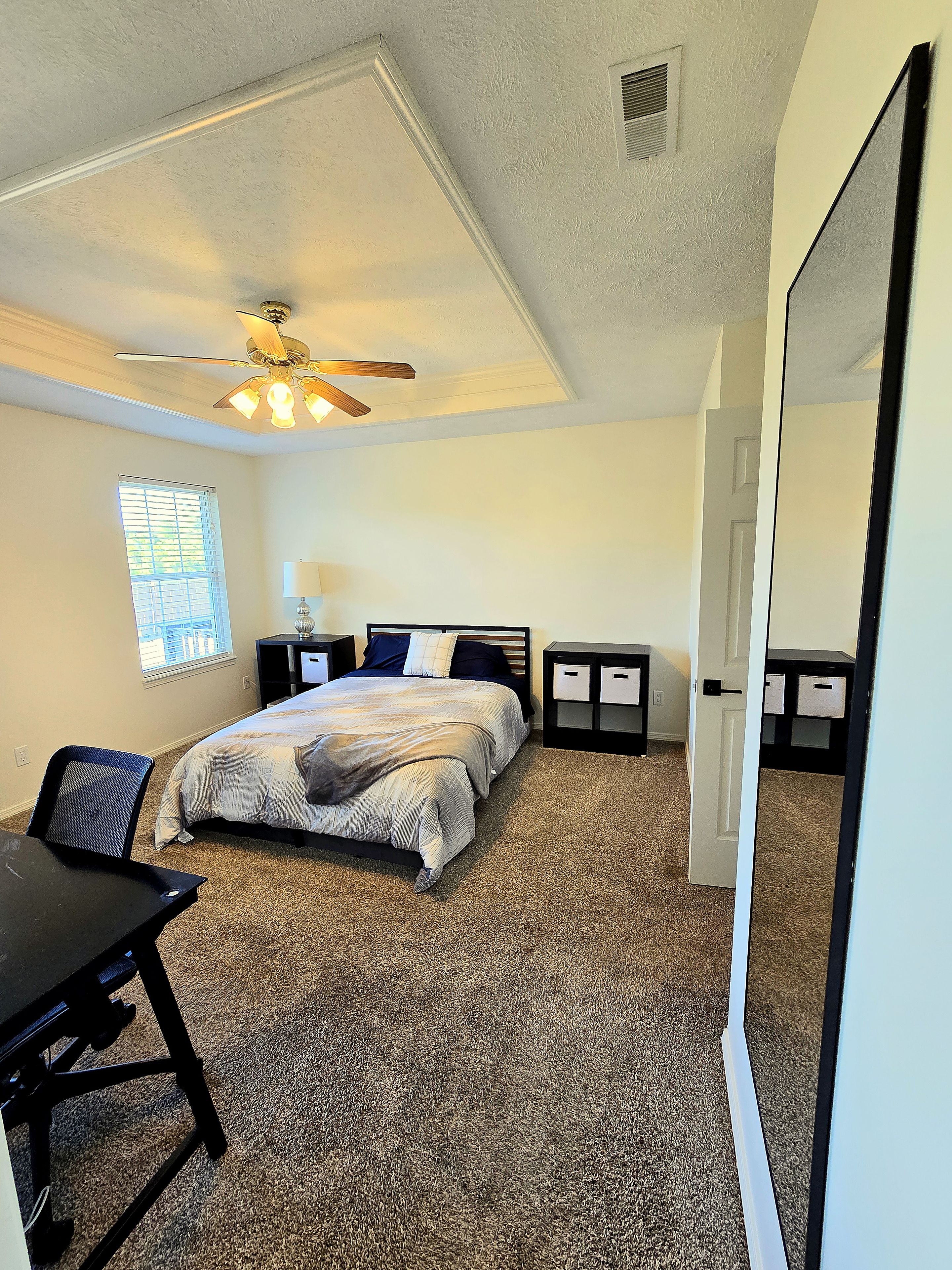 A bedroom with a bed, a desk, a chair, a ceiling fan, and a window.