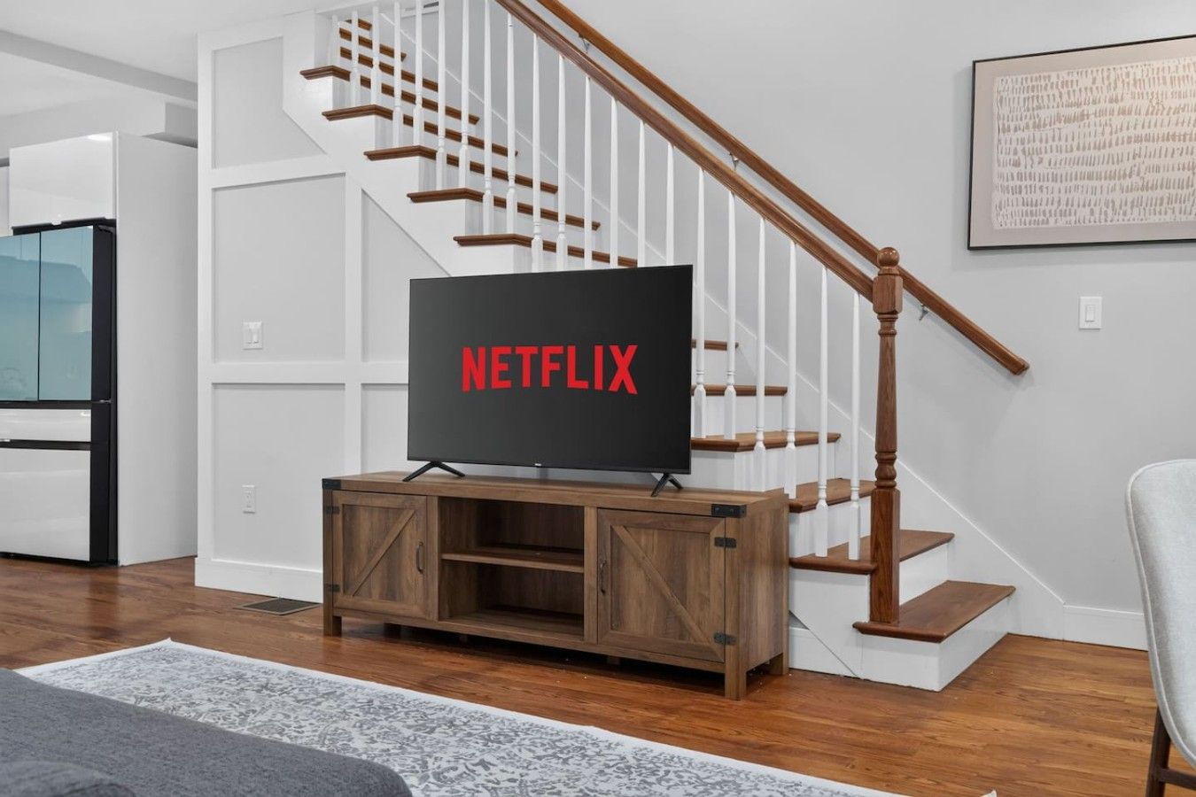 Un salon moderne avec un meuble de rangement en bois pour l’équipement audiovisuel, un téléviseur à écran plat affichant le logo « NETFLIX », et un escalier avec des rampes blanches.
