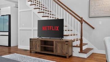 Un salon moderne avec un meuble de rangement en bois pour l’équipement audiovisuel, un téléviseur à écran plat affichant le logo « NETFLIX », et un escalier avec des rampes blanches.