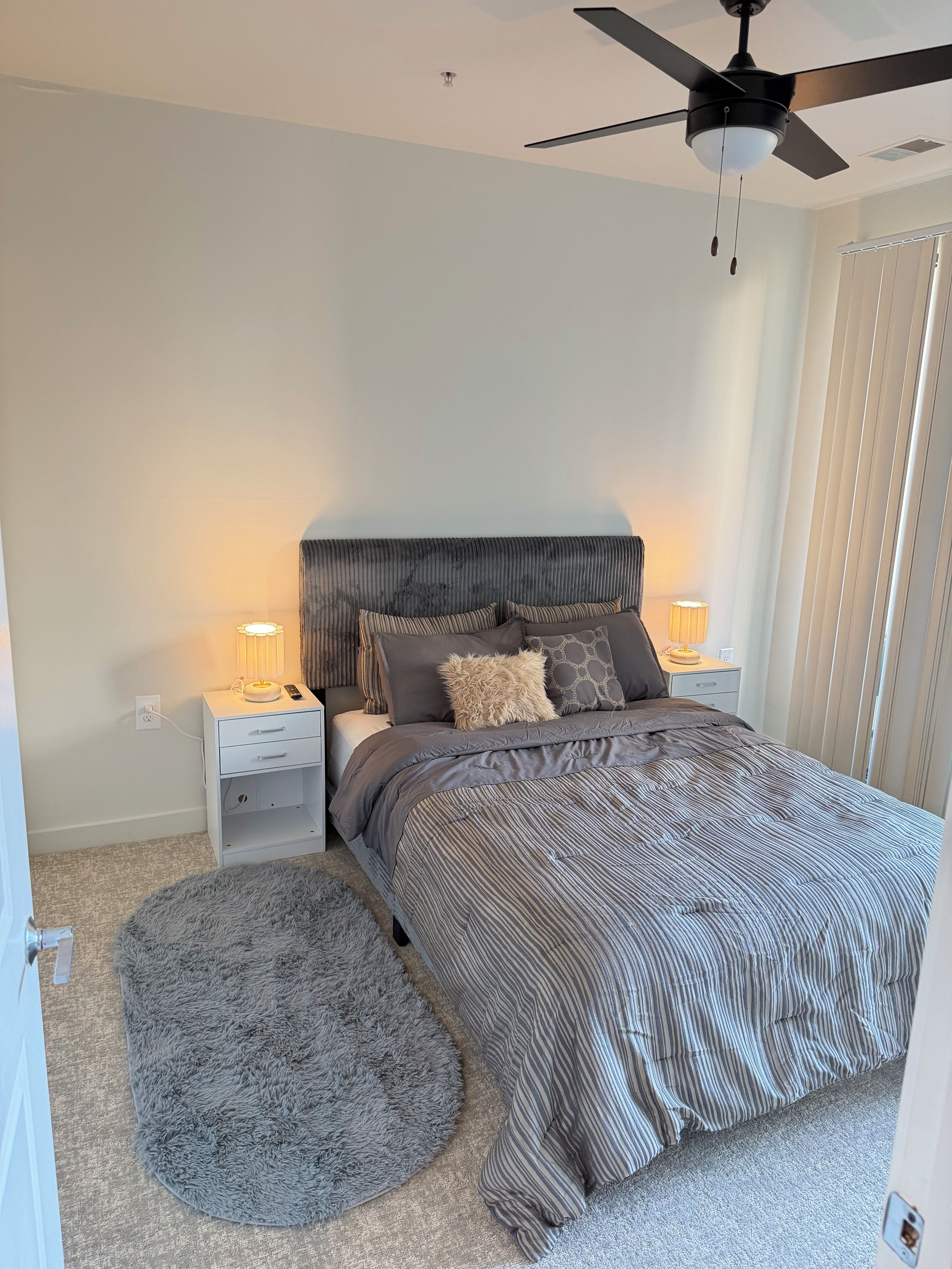 Un dormitorio con cama, mesitas de noche, alfombra redonda, ventilador de techo y ventana con persianas.