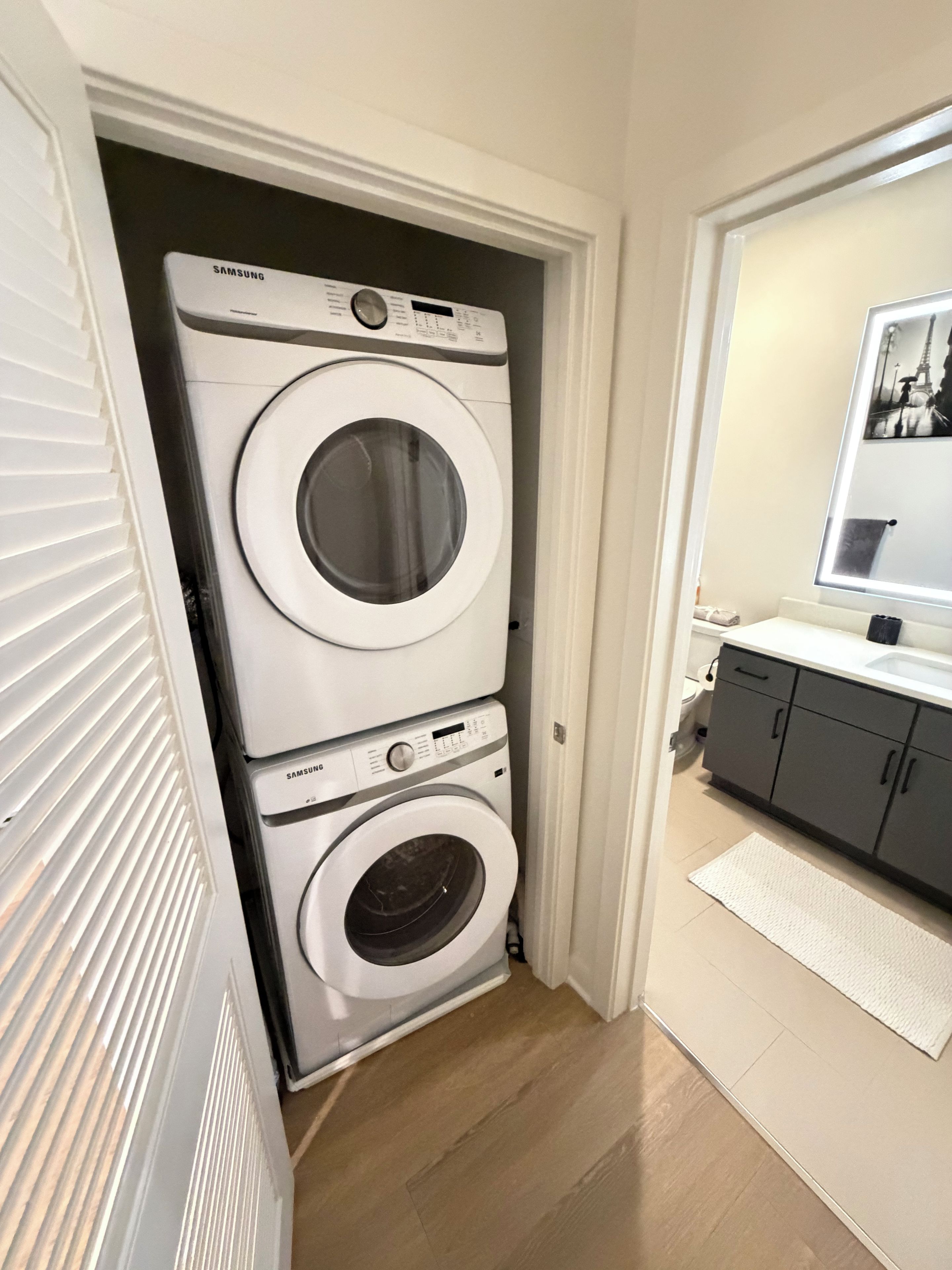 Un lavadero con dos lavadoras Samsung apiladas, un radiador blanco y una zona pequeña con lavamanos y espejo.