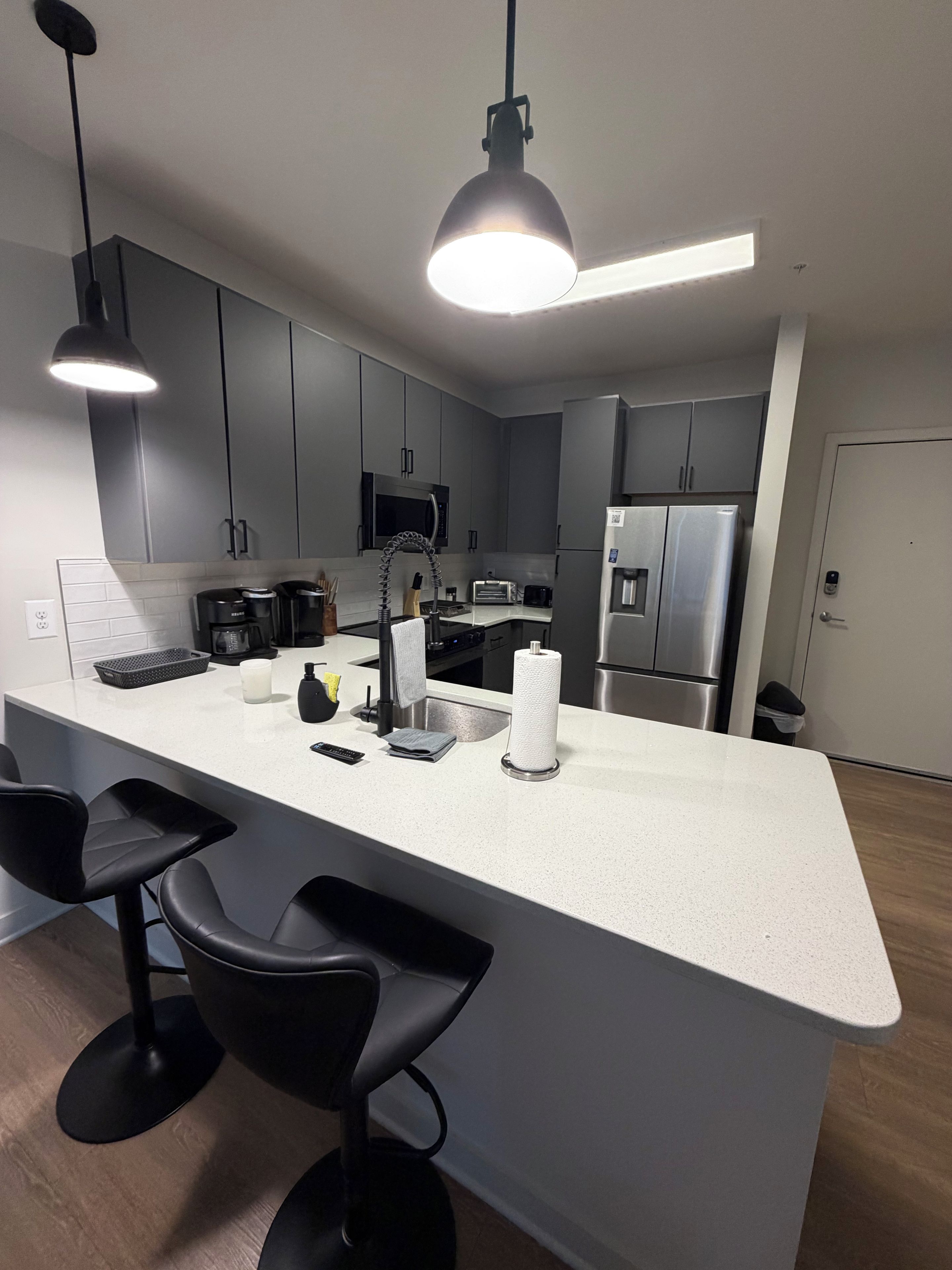 A modern kitchen with grey cabinets, a white countertop, a built-in microwave, a stainless steel refrigerator, and black bar stools.