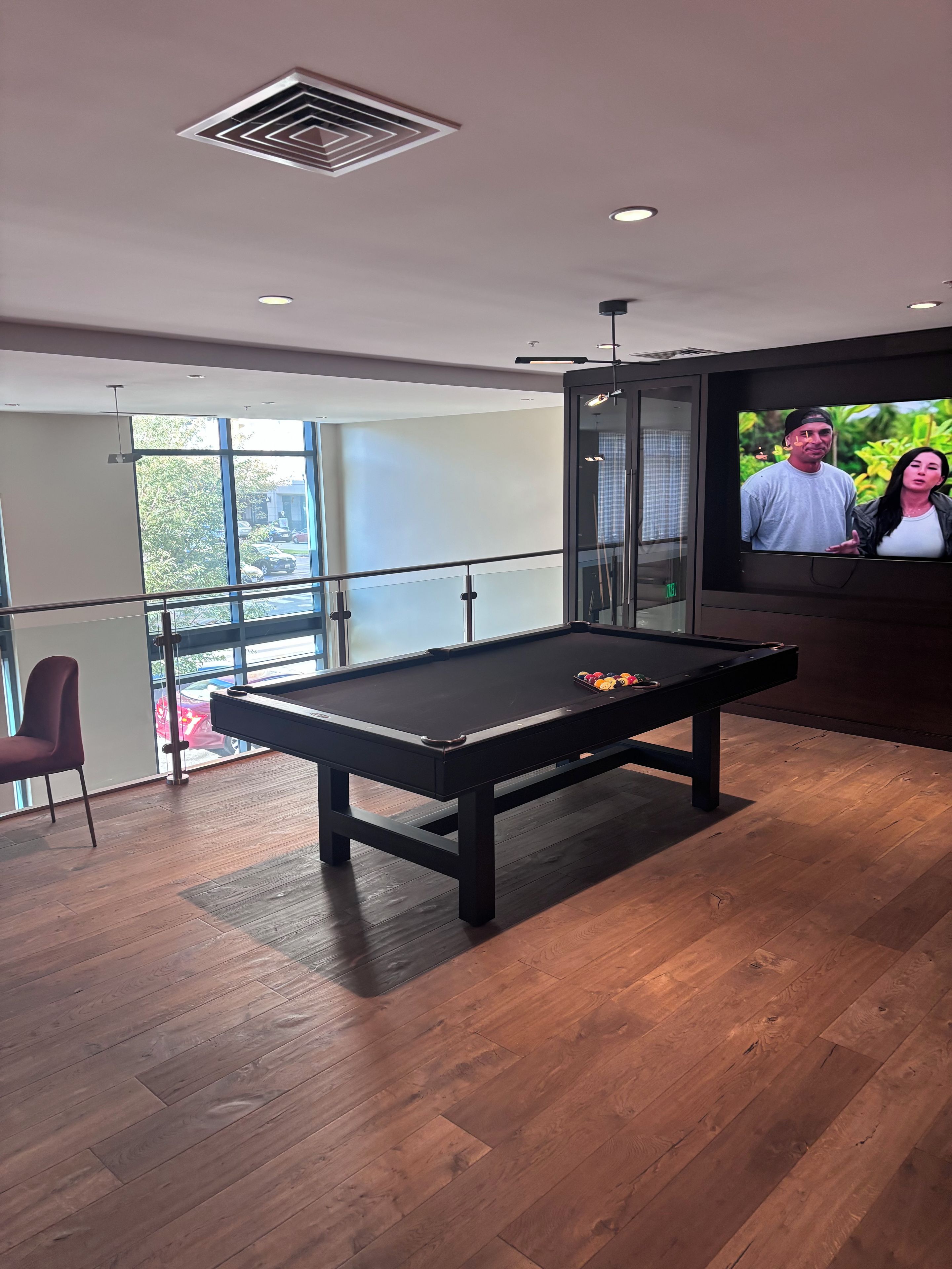 Une table de billard dans une pièce moderne, dotée d’une grande fenêtre, d’un téléviseur et d’une rambarde en verre.