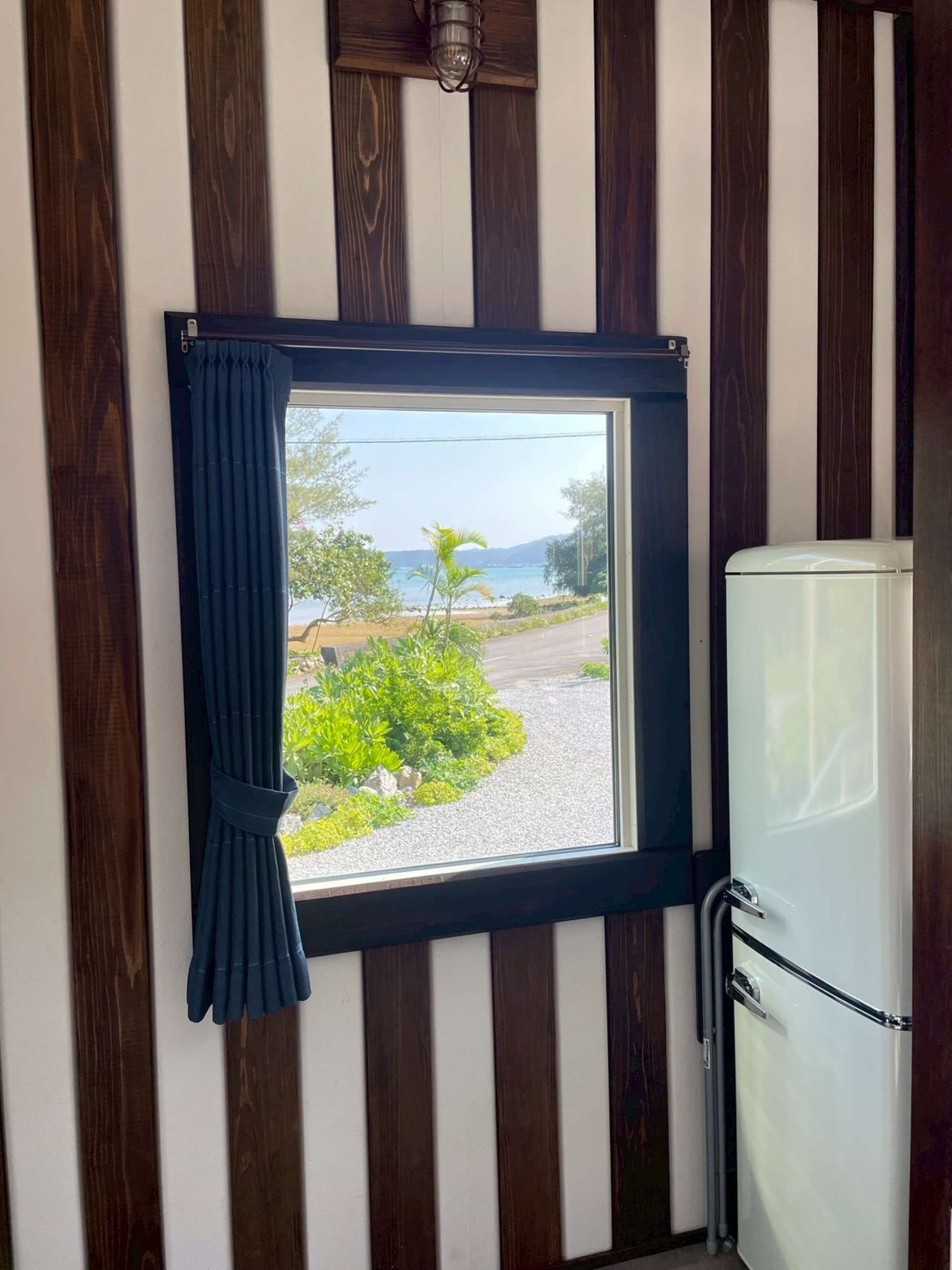 A window with a view of a beach and greenery, a white refrigerator, and wooden paneling on the walls.