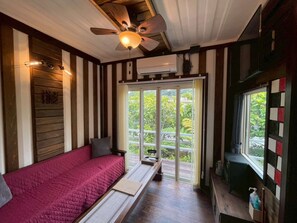 Una habitación con cama, ventilador de techo, ventana con vistas a vegetación y una mesita con una botella.