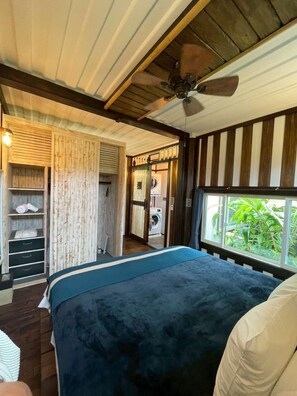 Un dormitorio con cama, ventilador de techo, ventana con vista a vegetación y armario de madera.