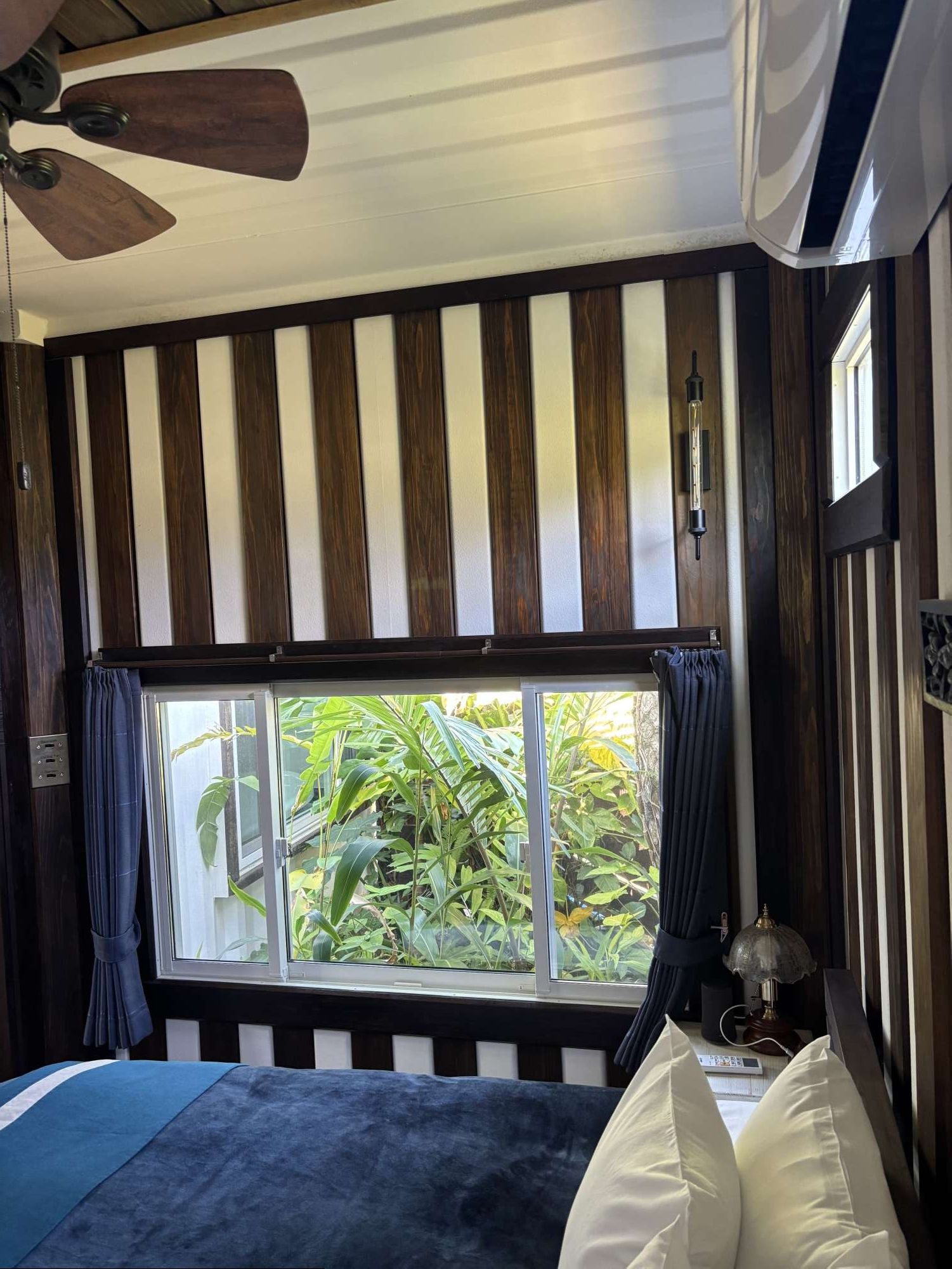 A room with a large window, a ceiling fan, and a bed with pillows.