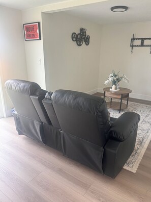 Un saló amb un sofà de cuir negre, una taula rodona petita amb un ram de flors, i un televisor fixat a la paret.