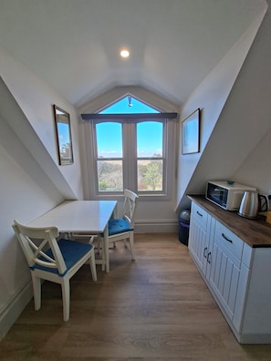 Une petite pièce mansardée triangulaire, comprenant une table et des chaises, un coin cuisine et une grande fenêtre donnant sur les arbres.
