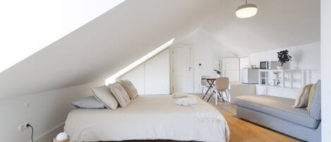 A modern loft bedroom with a sloped ceiling, a bed with pillows, a grey sofa, a small table, and a plant.
