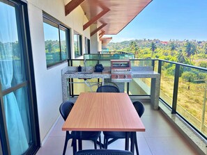 Un balcón con mesa y sillas de madera, una parrilla incorporada y vistas a un paisaje montañoso.