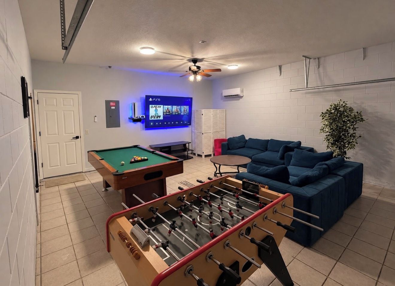 A room with a pool table, a foosball table, a blue sofa, and a television displaying a game.