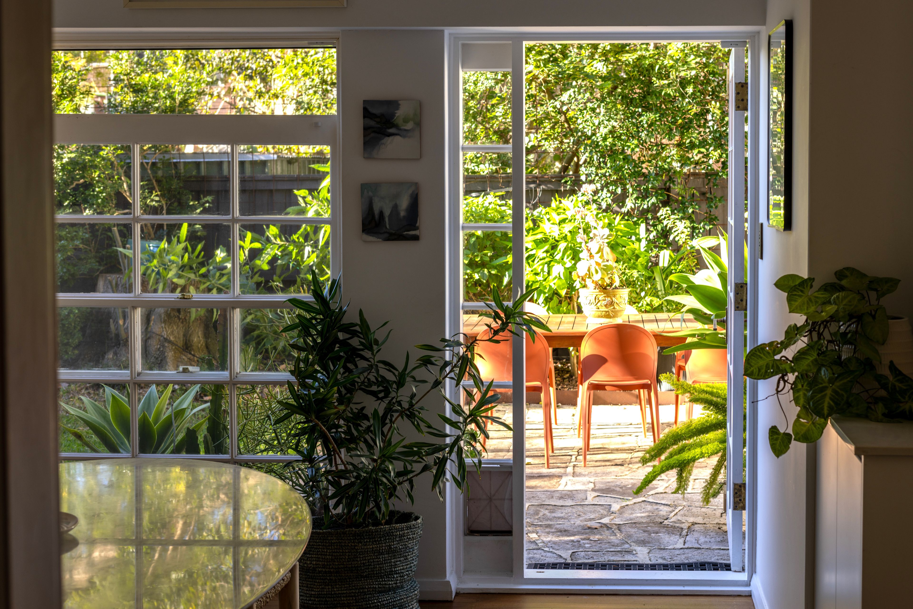A room with a glass door leading to an outdoor area with a table and chairs, surrounded by greenery.