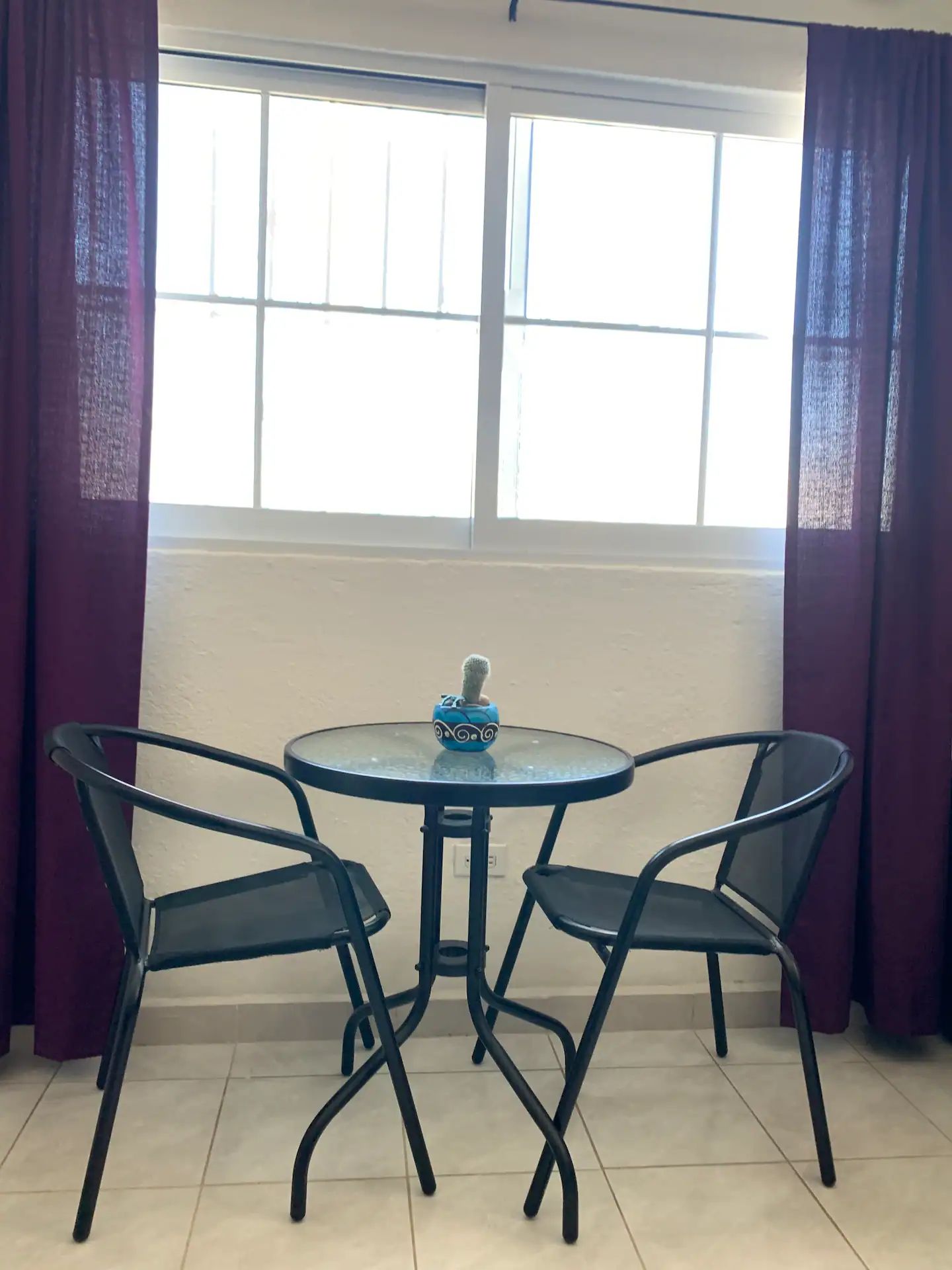 A small round table with two chairs, a cactus in a decorative pot, and a window with curtains.