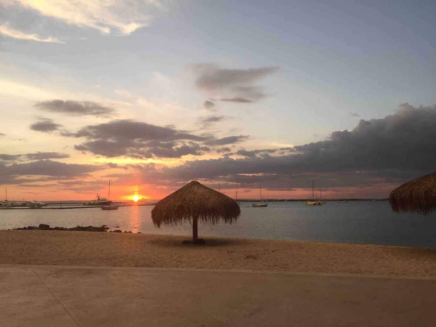 Een strandscène met een zonsondergang, een rieten hut en boten op het water.