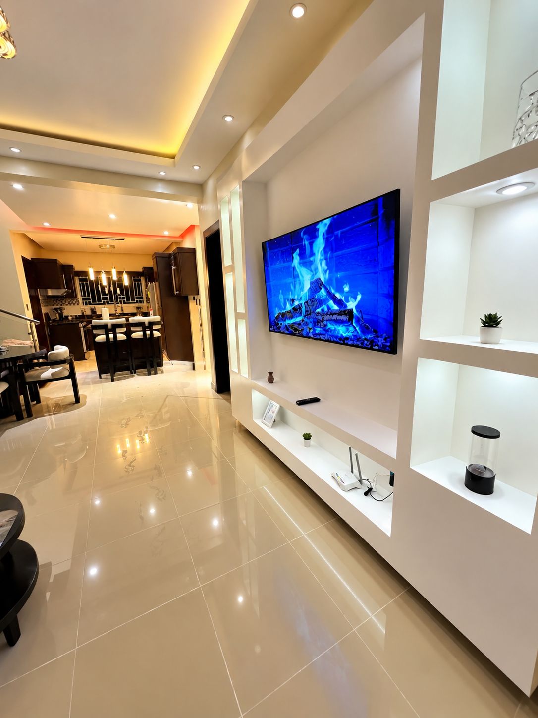 A modern living room with a flat-screen TV mounted on a white wall, a dining area with a table and chairs, and a kitchen in the background.