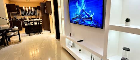 A modern living room with a flat-screen TV mounted on a white wall, a dining area with a table and chairs, and a kitchen in the background.