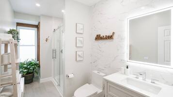 A modern bathroom with a glass shower enclosure, a toilet, a sink with a large mirror, and framed artwork on the wall.