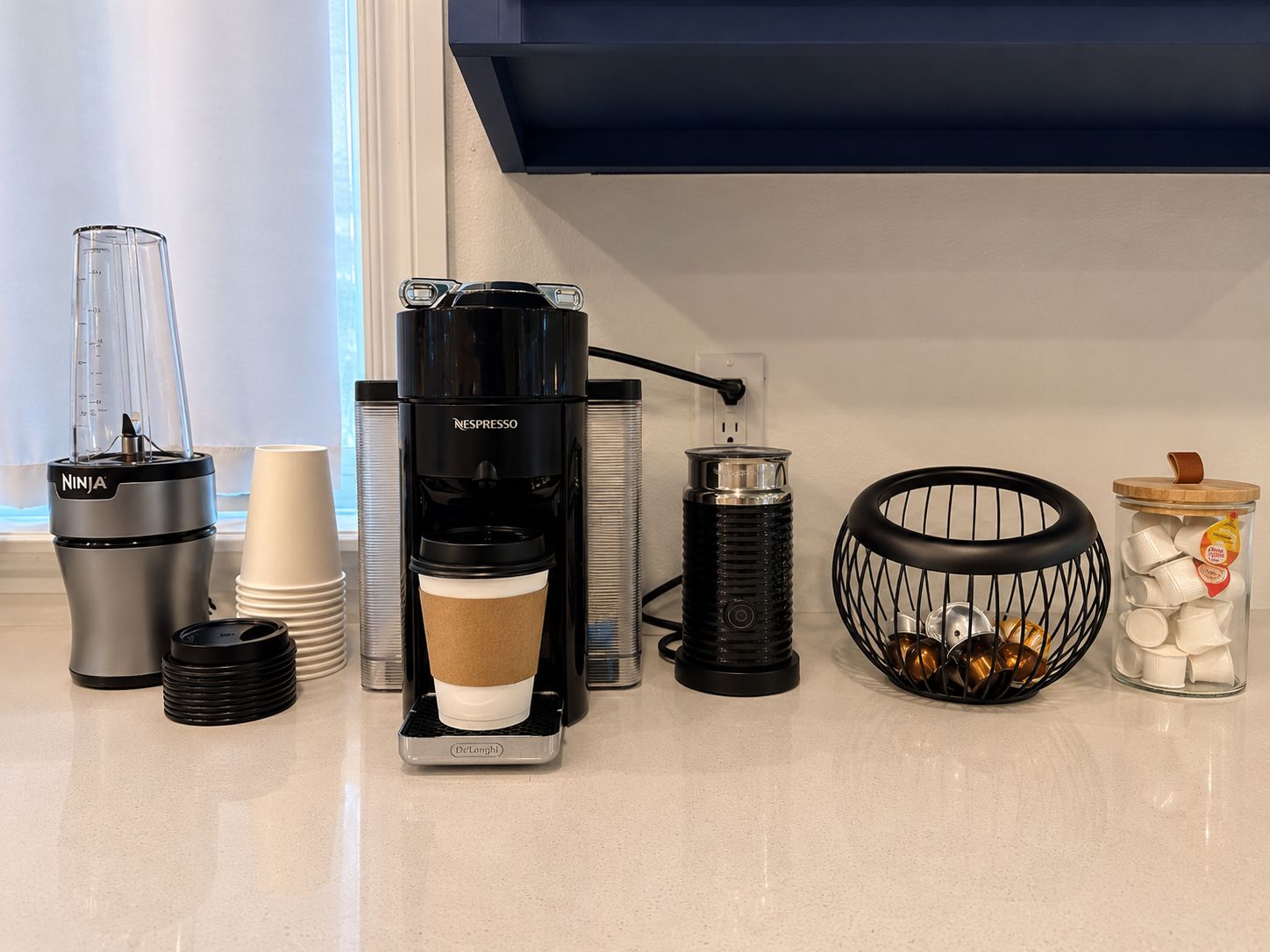 A kitchen counter with a coffee maker, a blender, a thermos, a coffee cup, a metal container, a candle holder, and a jar of sugar.