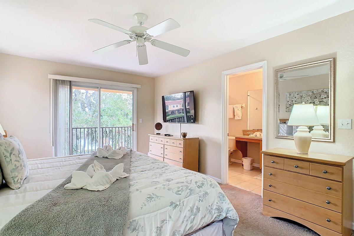 A bedroom with a bed, a dresser, a vanity, a ceiling fan, and a television.