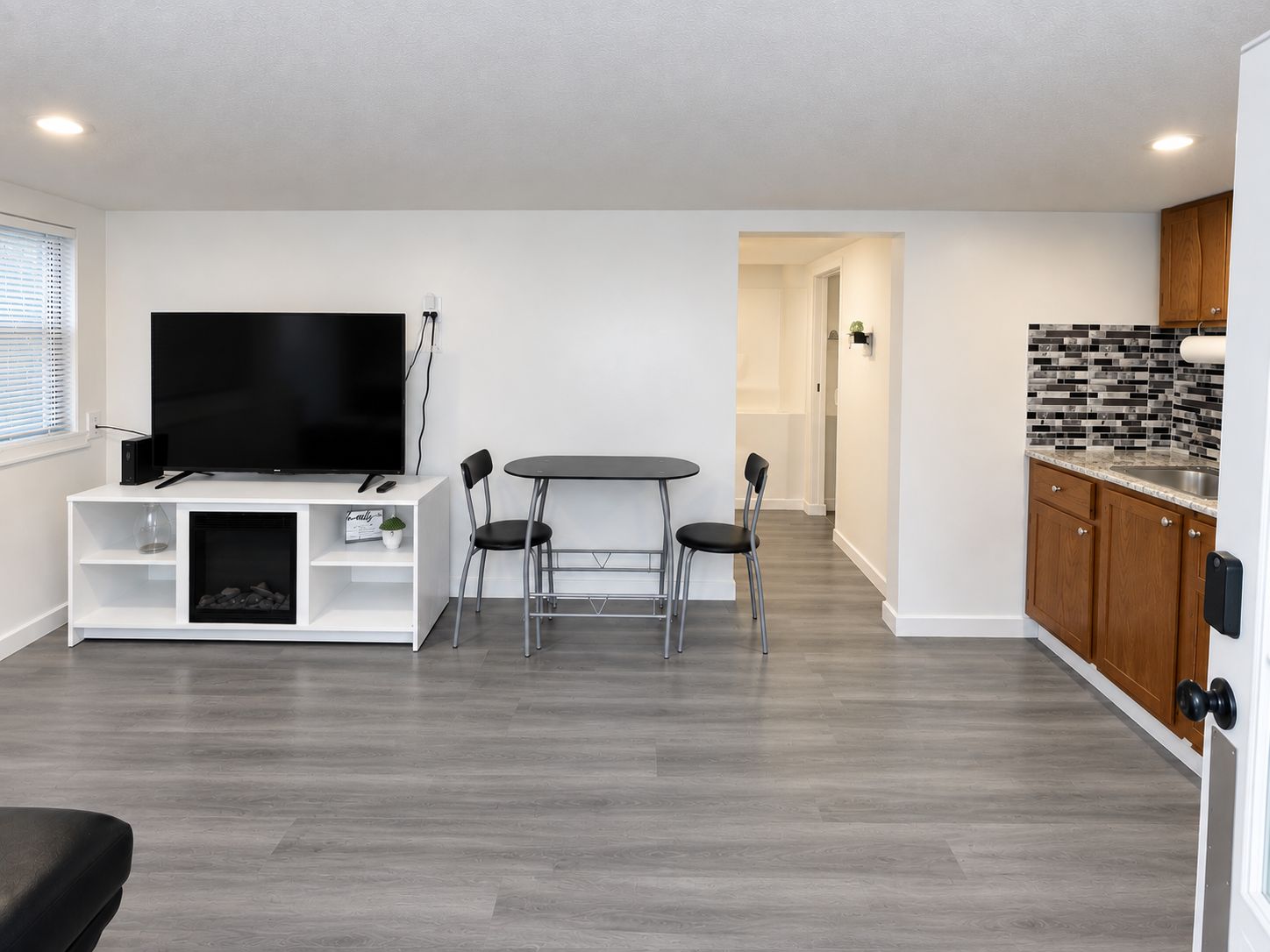 A modern living room with a flat-screen TV, a dining table with chairs, and a kitchen area with wooden cabinets and a tiled backsplash.