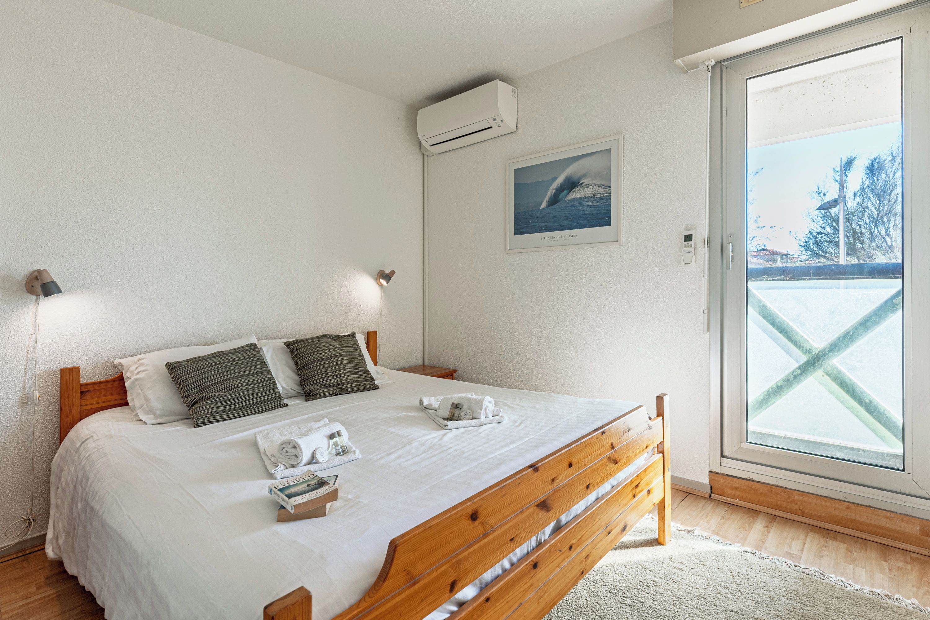 A bedroom with a wooden bed, white bedding, a mounted air conditioner, a framed picture, and a view through a glass door.