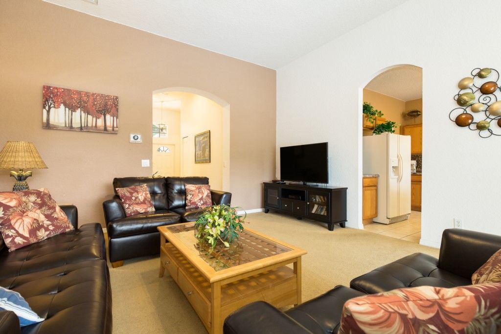A living room with a sofa set, a coffee table, a flat-screen TV, and a kitchen visible through an archway.