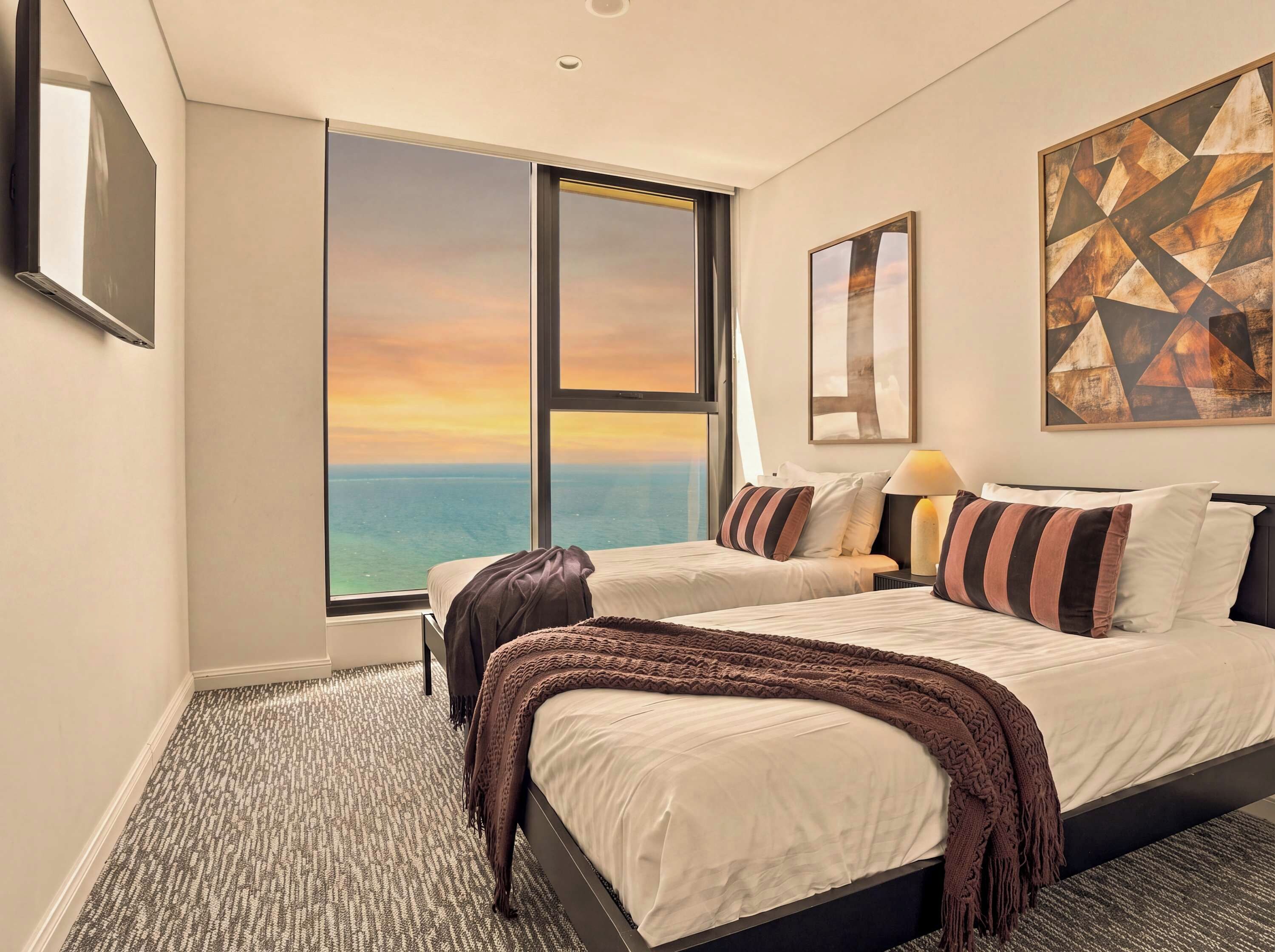 Une chambre d’hôtel dotée d’une grande fenêtre donnant sur la mer, d’un lit avec une tête de lit sombre et d’un tableau accroché au mur.
