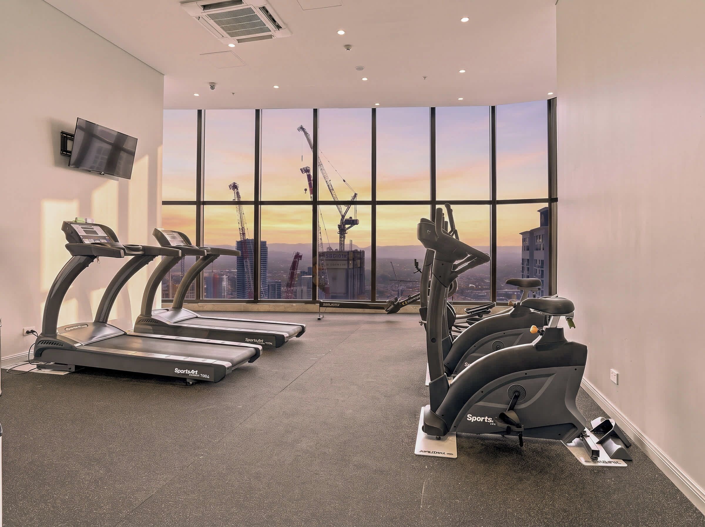 Ein Fitnessstudio mit Laufbändern und Ergometer, große Fenster mit Blick auf die Stadt sowie ein an der Wand befestigter Flachbildfernseher.