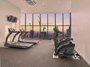 A gym with treadmills and stationary bikes, large windows with a cityscape view, and a flat-screen TV mounted on the wall. - The Mojo Sky Suite, Sunrise Ocean View, Meriton (Surfers Paradise)