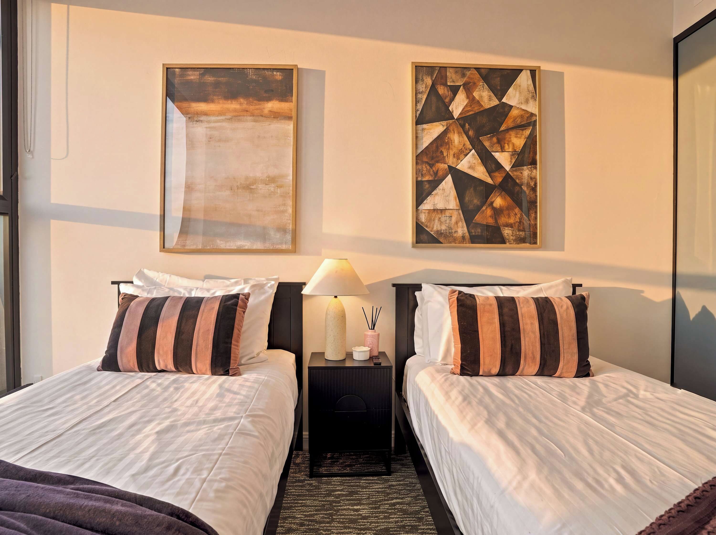 Une chambre d’hôtel avec deux lits, une table de chevet avec une lampe et deux tableaux abstraits accrochés au mur.