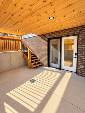 Un espace extérieur abrité, doté d’un escalier en bois menant à un mur en briques, d’une porte-fenêtre coulissante et d’un sol en béton.