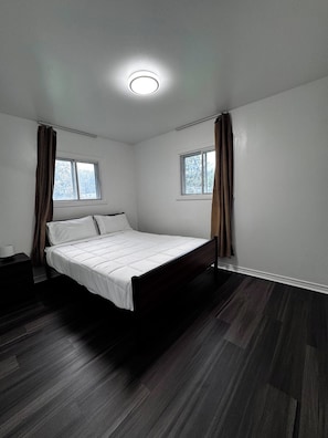 Une chambre à coucher avec un lit, deux fenêtres, une table de chevet et un plancher en bois foncé.