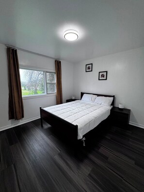 Une chambre à coucher avec un lit, une fenêtre avec des rideaux, une table de chevet et un luminaire fixé au mur.