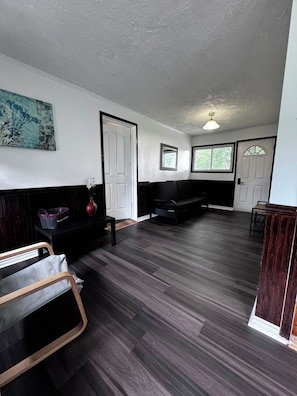 Une pièce avec un parquet en bois foncé, un canapé noir, une chaise en bois, une petite table et un tableau accroché au mur.