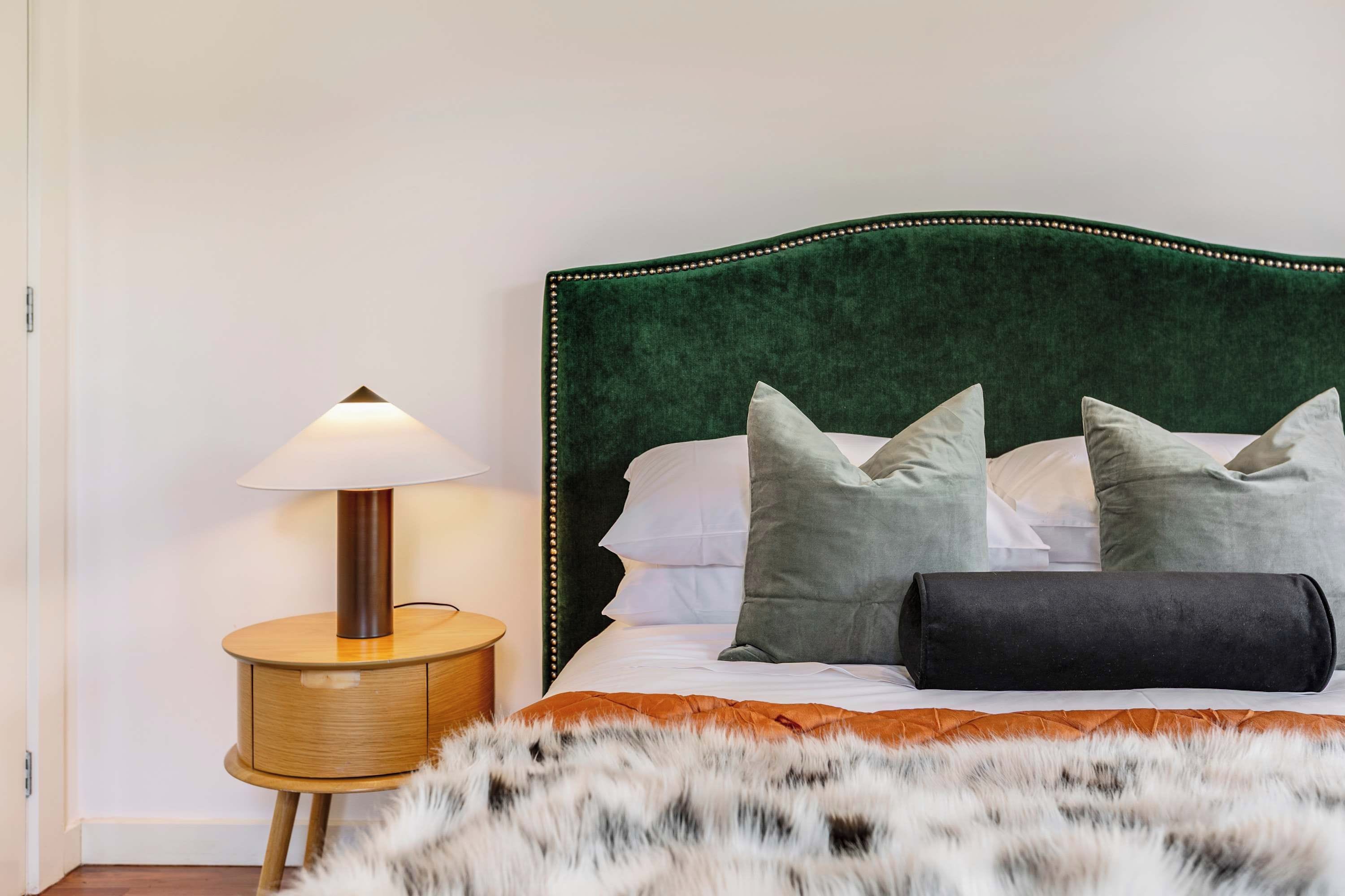 A bedroom with a green upholstered headboard, a wooden bedside table with a lamp, and a fur throw on the bed.