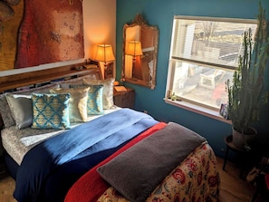 Une chambre à coucher avec un lit, des oreillers, une table de chevet, un miroir, une fenêtre avec des stores et une plante en pot.